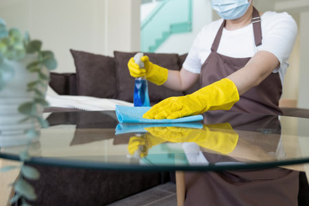 Young beautiful female housekeeper, housewife wearing apron, protective gloves, and face mask standing at table holding alcohol spray and cloth to clean and wipe table in living room at home. Cleaning, housekeeping concept.の写真素材