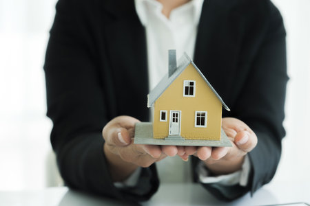 Female real estate agent in suit is sitting at table with contract document and holding house model and is handing property keys to her customer displaying succesful negotiation and decision to invest and purchase a property.の写真素材