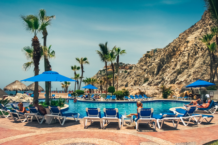 Cabo San Lucas / Mexico - August 13, 2007: View on the hotel pool with people sunbathing on the blue sunbeds. Rocky hill and the ocean in the background.のeditorial素材