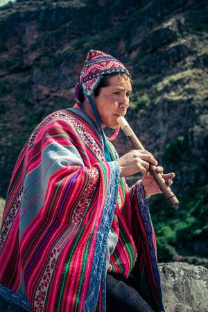 Cusco / Peru - May 29.2008: Portrait of a man, shepherd, goat herder, dressed up in native, peruvian costume seating on the rock and playing on the flute in the mountains.