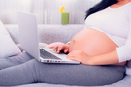 Pregnant woman using her laptop while relaxing on the couch at home and exposing her pregnant belly.