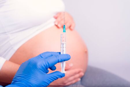 Pregnant woman having medicine injection. Hand with blue gloves holding syringe in front of the pregnant belly.の写真素材