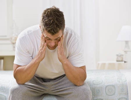 A man is seated on the bed in a bedroom. He has his head in his hands and is looking away from the camera. Horizontally framed shot. A man is seated on the bed in a bedroom. He has his head in his hands and is looking away from the camera. Horizontally framed shot.