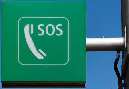 Close up of metal arm mounted green SOS sign and telephone symbol indicating emergency call facility in a public place against blue sky backgroundの写真素材