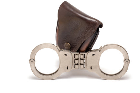 Close up police iron metal steel handcuffs with rigid intermediate piece in front of dark leather case closed wrist locking bars on white background as concept for arrest and imprisonmentの写真素材