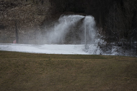Snow lance cannon producing artificial snow on slopes and in ski resorts with green brownish grass in foreground during winter with missing snow as a concept for global warming and climate changeの写真素材