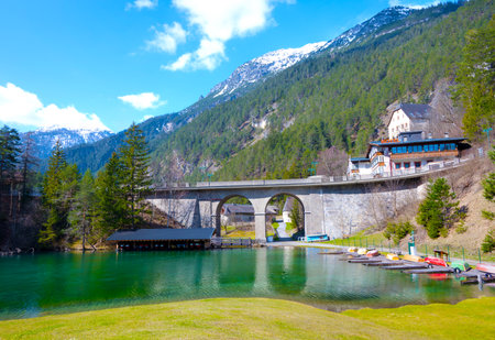 Landscape shot of clear reflecting mountain lake with jetty rowing boats boathouse and high road bridge with two round arches viaduct in front of high mountains and partly cloudy blue sky in Tyrolの写真素材