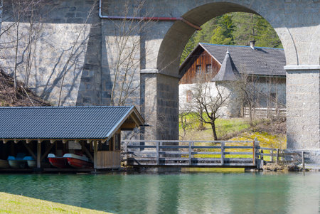 clear reflecting mountain lake with wooden bridge boathouse pedal boats and an old tower framed in a round arch of a massive stone bridge with farmhouse in Tyrolの写真素材