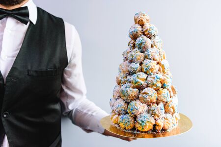 Crockenbush or Profiterole tower (cone of glazed profitroles) on blue background in server's hands....