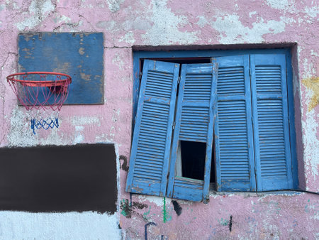 Old Mediterranean house exterior wall. Abandoned building with cracked facade, blue broken window shutters and basketball hoop, old crumbling plaster. Athens, Greece.の写真素材