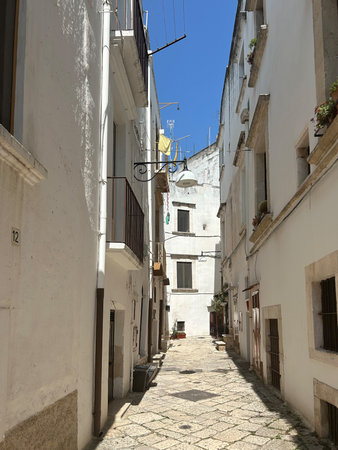 A narrow street lined with old stone buildings and balconies winds through a quiet European town. Potted plants, shuttered windows, and warm sunlight create a charmingの写真素材