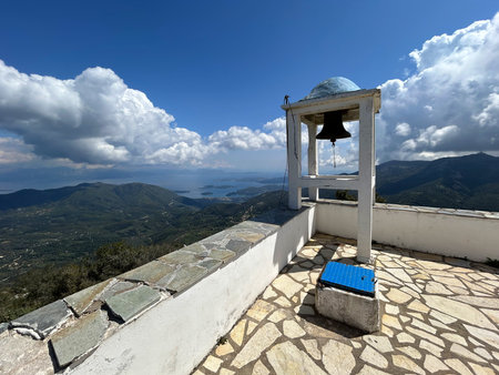 A bell tower stands on a mountain terrace overlooking green hills and the blue sea. The sky is covered with puffy clouds, casting shadows. The view is vast, peacefulの写真素材