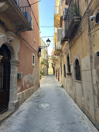 A narrow street lined with old stone buildings and balconies winds through a quiet European town. Potted plants, shuttered windows, and warm sunlight create a charmingの写真素材
