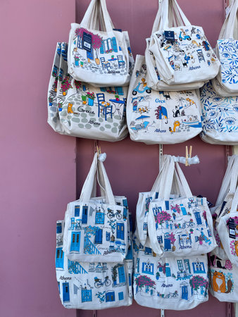 Colorful fabric tote bags with Greece and Athens illustrations hanging on a pink wall. Souvenir cotton bags featuring traditional blue doors, cats, and Mediterranean street scenes.の写真素材