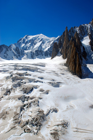 Alps mountains covered in ice and snow with cracks on high altitude, with summit in the background, black rocky sharp pokes and clear blue skyの写真素材