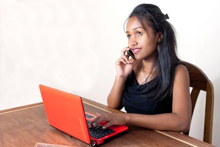 A young woman working at home on her laptop computer while talking on a mobile phoneの写真素材