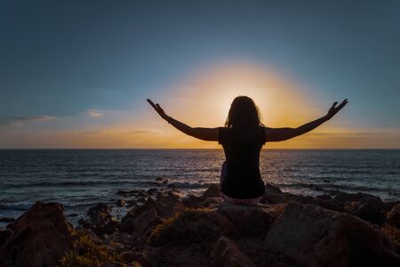 A woman praticing the lotus posture of Hatha yoga at sunset in front of the beach