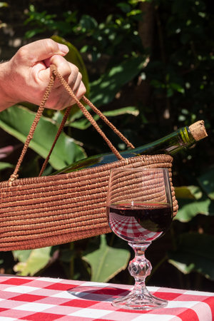 Hand holding a bottle of red wine with a straw bag in the backgroundの写真素材