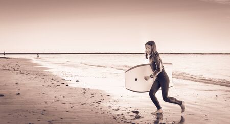 A girl dressed in a wetsuit runs on the beach with a bodyboard
