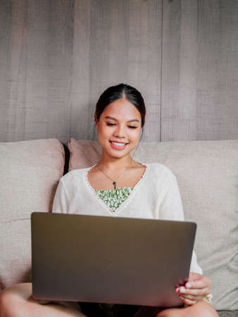 Beautiful young smiling asian woman working on laptop in living room at home. Asia business woman working in her home office. Enjoying time at home.の写真素材