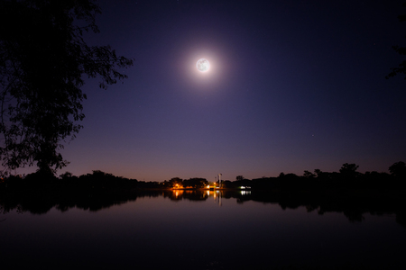 Full moon shine bright in the night above the big lake and small villageの写真素材