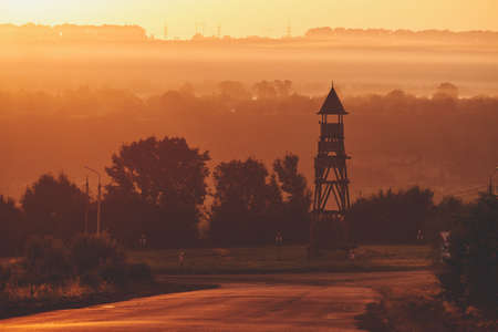 Foggy evening at sunset. Bluish fog in the sun. Ancient wooden tower in the middle of the road ring. Asphalt road in the fog. Electric poles on the horizon.の写真素材