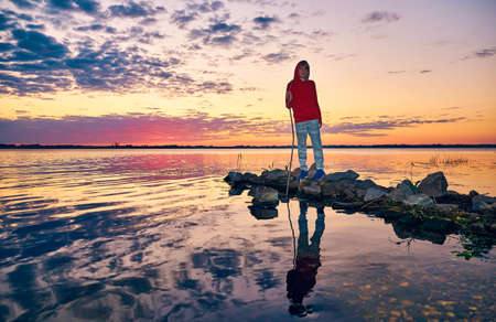 A teenager in a red hoodie and gray pants and blue sneakers stands on the stone pier of the river....