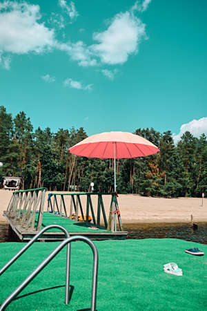 There is a pink beach umbrella on the green pier. Sunny weekend. Pines can be seen on the river bank.の写真素材