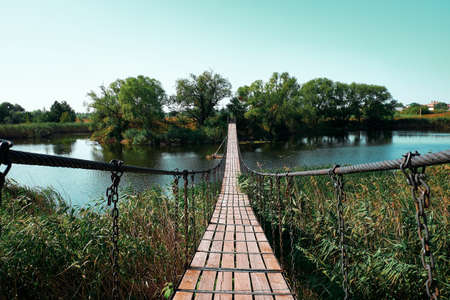 Suspension bridge across the river to the island with deciduous trees. Wooden battens are fastened...