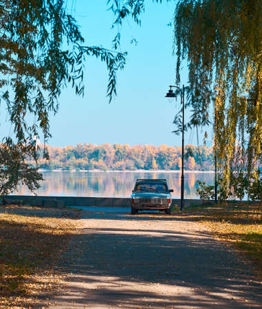 An old car stands under a street lamp. Autumn on the river. The trail is covered with fallen willow...