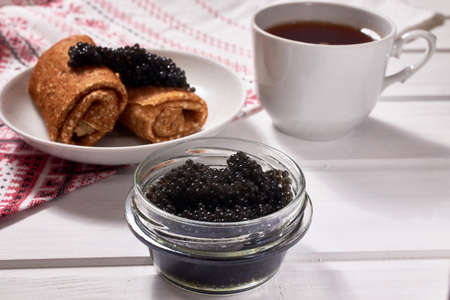 A glass jar of black caviar on a white wooden background. Traditional food for Maslenitsa