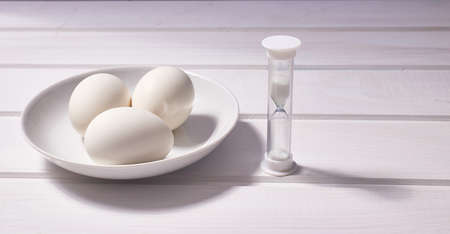 Three eggs and an hourglass on a white wooden background. Copy space
