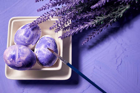 Coloring eggs with a brush. Easter tradition. Painted eggs on a purple watercolor background and purple lavender flowers. Selective focus. With copy spaceの写真素材