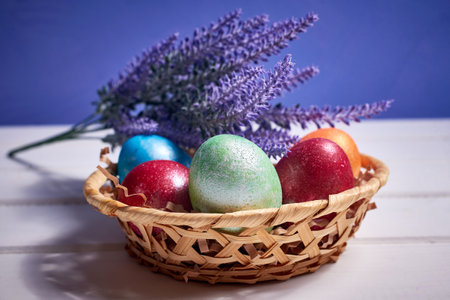 A small basket with colorful painted eggs and a bouquet of lavender on a white wooden background....