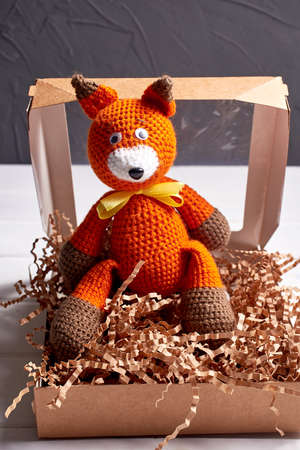 A beautiful knitted fox toy in a brown cardboard box on a gray background. Selective focus.の写真素材
