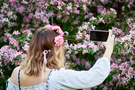 A young girl in a wreath of roses takes a selfie in the background of a flowering tree. An idea for...