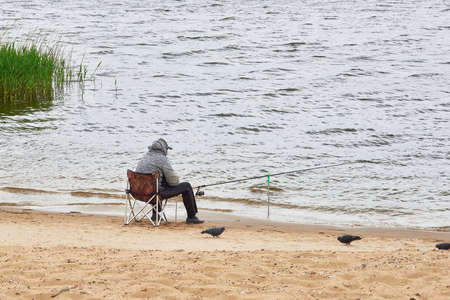 A fisherman sits on a chair and catches fish with a fishing rod in the river. The pigeons on the sand look at him.の写真素材