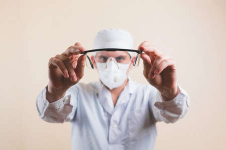 Protective glasses in doctors hands in white lab coat and protective breathing mask defocused on...