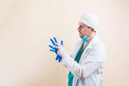 Young doctor in white suit and protective breathing mask puts gloves on his hands on yellow...