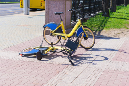 Two vibrant yellow rental bicycles on a sidewalk, one of them toppled on its side by vandals. In the background, a yellow bus is seen traveling on the road.の写真素材