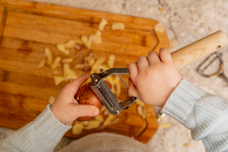 Child's Hands Using a Metal Vegetable Peeler to Peel a Potato Over a Wooden Cutting Boardの写真素材