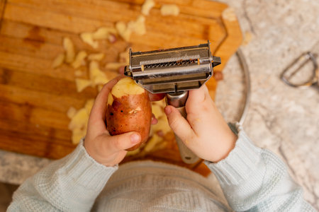 Child's Hands Using a Metal Vegetable Peeler to Peel a Potato Over a Wooden Cutting Board