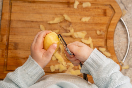 Child's Hands Peeling a Potato Over a Wooden Cutting Board with a Metal Vegetable Peeler