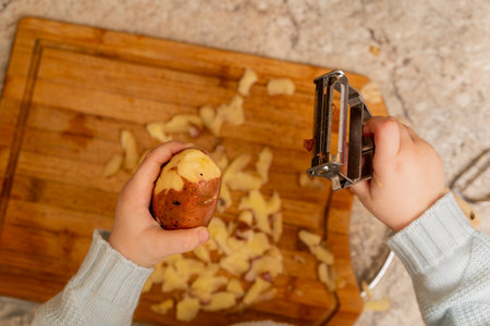Child's Hands Using a Metal Vegetable Peeler to Peel a Potato Over a Wooden Cutting Boardの写真素材