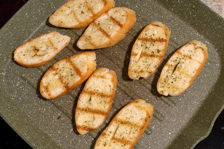 Several slices of seasoned and grilled bread on a speckled non-stick grill pan. The bread slices have visible grill marks and herbs, possibly garlic.の写真素材