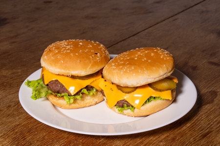 Two cheeseburgers on a white plate, featuring sesame seed buns, beef patties, melted cheese slices, pickles, and crisp lettuce, set on a rustic wooden surface.の写真素材