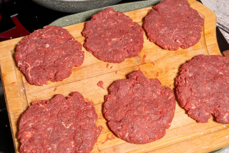 Six uncooked hamburger patties made of ground meat are placed on a wooden cutting board. In the background, hamburger buns are being toasted in a frying pan, illustrating burger preparation.の写真素材