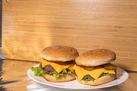 Two cheeseburgers with sesame seed buns, beef patties, cheddar cheese, pickles, tomato slices, and lettuce are served on a white plate. A rustic wooden surface forms the background.の写真素材