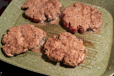 Four hamburger patties cooking on a green speckled griddle. The patties show visible browning and texture, surrounded by grease, emphasizing the sizzling preparation process.の写真素材