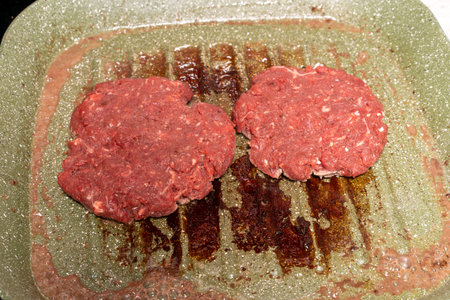Two raw hamburger patties rest on a speckled green grill pan with visible grease marks. The coarse-textured patties highlight the preparation stage of homemade burgers, ready for grilling.の写真素材